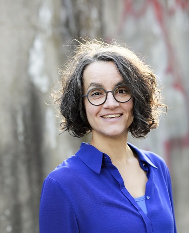Saskia Bellem, kinnlange braune Locken, dunkle runde Brille, kobaltblaue Bluse, steht draußen vor grauer Mauer. (c) Marlène Meyer-Dunker