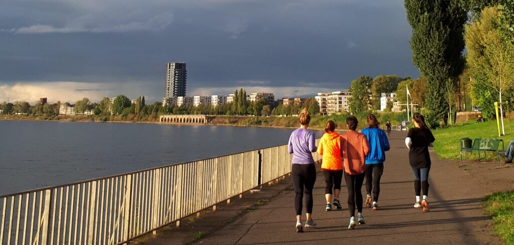 eine Gruppe Frauen in farbigen Sportsachen laufen draußen am Rhein, hinten dunkle Wolken